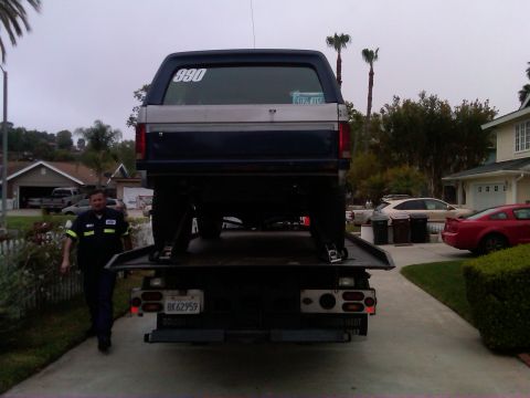 1986 Ford Bronco - A tow truck driver stands beside a Ford Bronco being loaded onto a flatbed truck in a residential area.