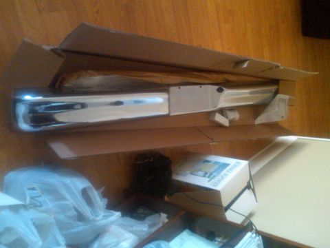 1986 Ford Bronco - A chrome bumper is shown partially unpacked from its cardboard shipping box, resting on a wooden floor.