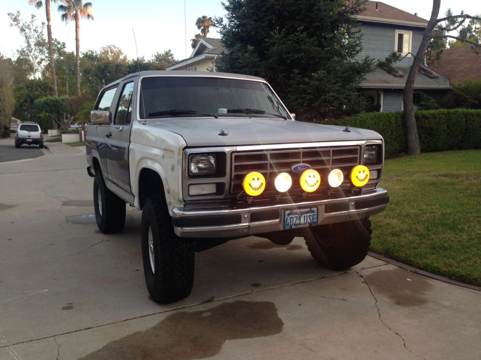 Gray and white Ford Bronco with smiley face fog lights and off-road tires parked in a driveway