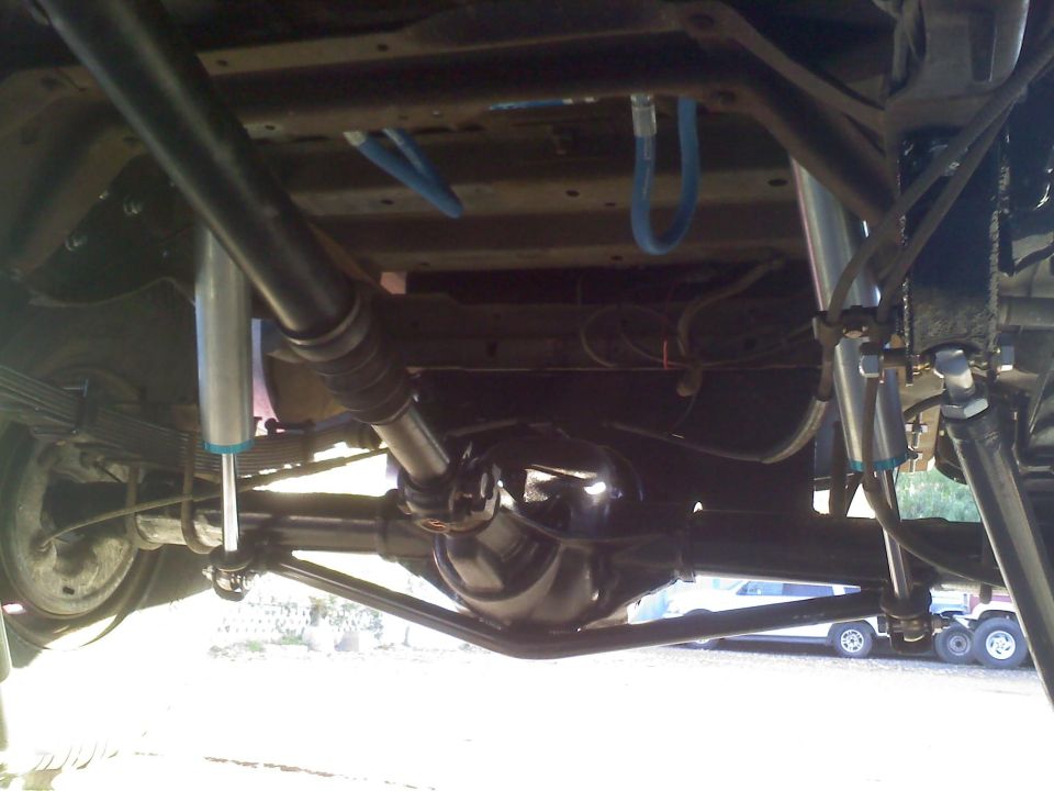 Underside view of a vehicle's rear axle, driveshaft, and suspension with blue hoses. Another vehicle is visible in the background