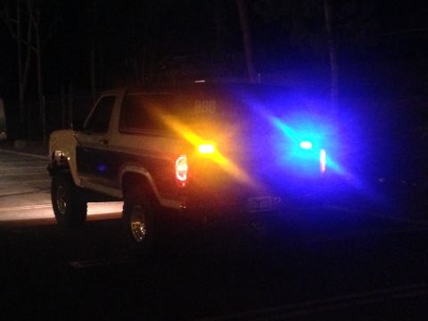 1986 Ford Bronco - A Ford Bronco is seen from the rear at night, illuminated by flashing yellow and blue emergency lights.