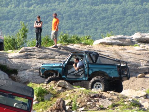 1979 Ford Bronco - A teal off-road SUV with a roll cage is seen navigating a rocky trail, with two people standing on a ledge above.