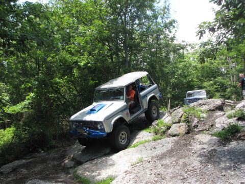 1979 Ford Bronco - Two Ford Broncos navigate a challenging rocky terrain on a wooded trail.