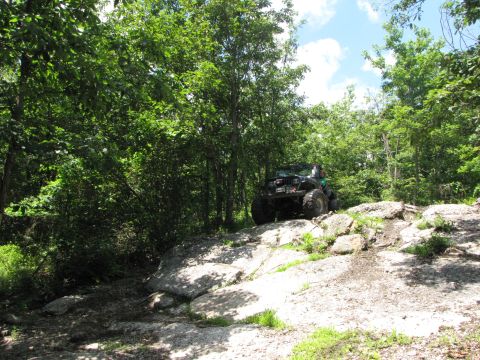 1979 Ford Bronco - An off-road vehicle navigates a rocky incline on a wooded trail under a partly cloudy sky.