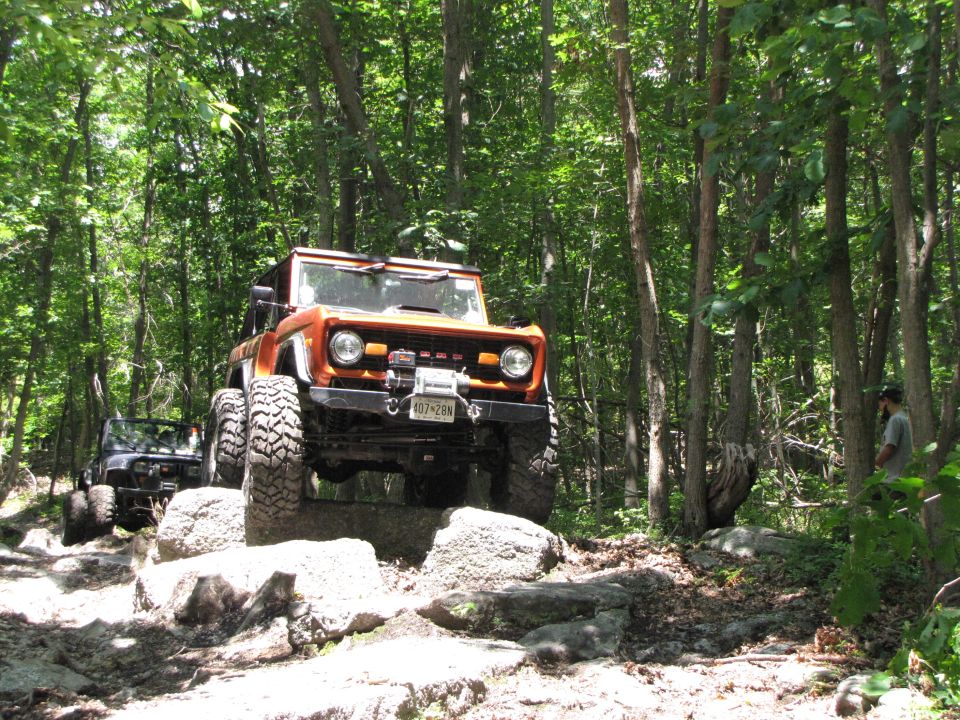 orange 1979 Ford Bronco climbing rocks on wooded trail, another SUV behind it