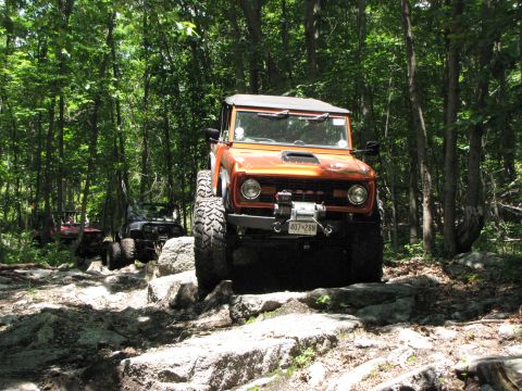 1979 Ford Bronco - An orange 1979 Ford Bronco, modified for off-roading, tackles a challenging rocky path in a wooded area, with other vehicles following behi…