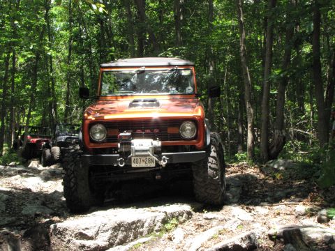 1979 Ford Bronco - An orange Ford Bronco navigates a rocky path through a wooded area, with another vehicle following behind.