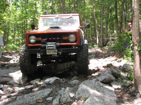 1979 Ford Bronco - An orange Ford Bronco with oversized tires is shown on a challenging, rocky off-road path.