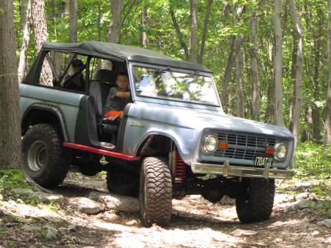 1979 Ford Bronco - This light blue off-road SUV navigates a challenging rocky trail through the woods, its soft top providing open-air enjoyment.