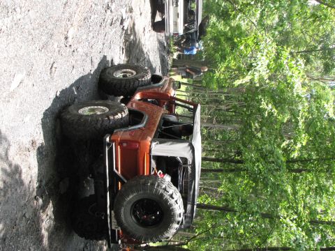 1979 Ford Bronco - An orange off-road vehicle navigates a rocky path surrounded by trees, with another vehicle visible in the background.
