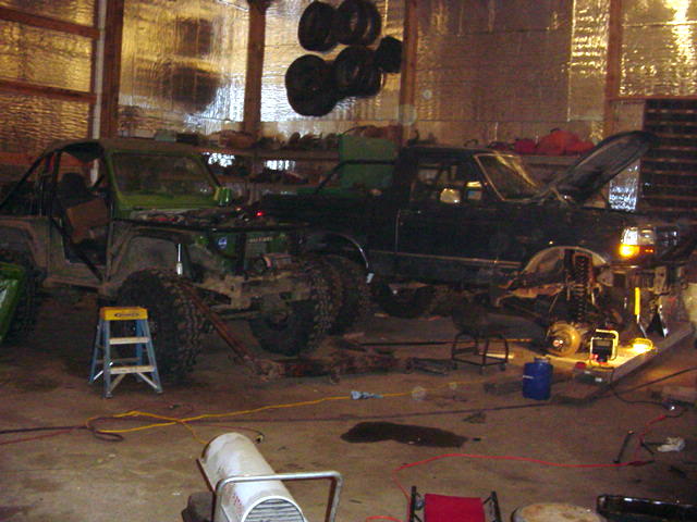 Two off-road vehicles, one green and one black, are in a garage with the black vehicle's hood open and engine exposed