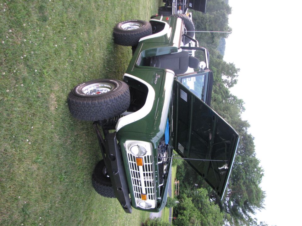 Green Ford Bronco with white stripe, hood open, parked on grass. Another vehicle is visible behind it