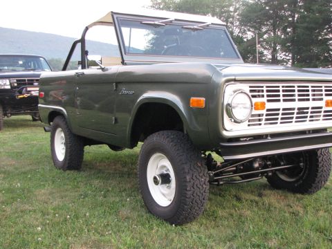 1979 Ford Bronco - A classic green Ford Bronco is shown from the front quarter angle, with a second vehicle partially visible behind it.