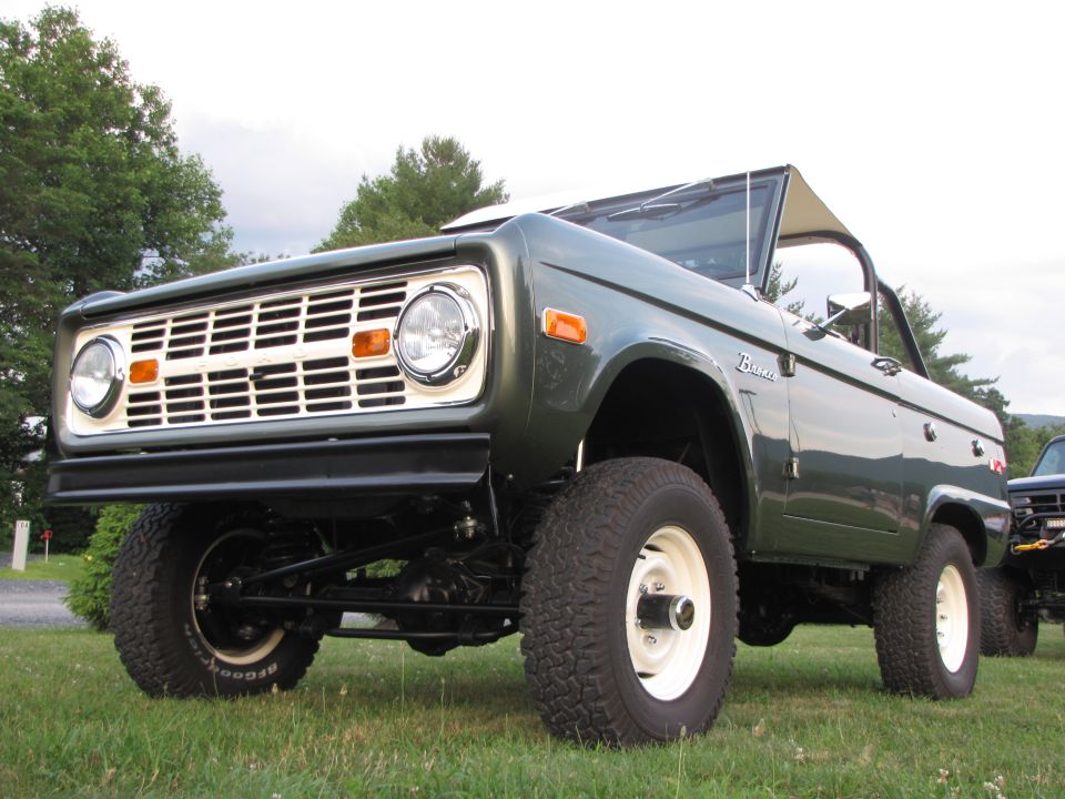 Green Ford Bronco with white grille and white wheels, parked on grass. Another vehicle is partially visible to the right
