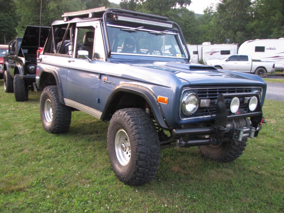 Blue Ford Bronco with aftermarket wheels, tires, winch, and light bar. Another black Bronco is parked behind it