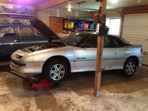 1990 Geo Storm GSi - A silver coupe is being worked on in a garage, with a blue car parked behind it.
