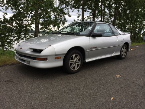 1990 Geo Storm GSi - A silver coupe with a black roof is parked on an asphalt surface near a body of water and trees.