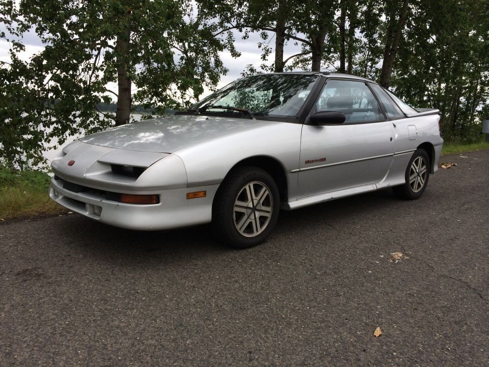 Silver Isuzu Impulse coupe parked by a lake with trees