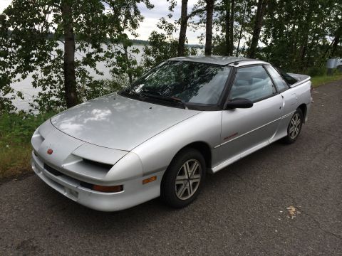 1990 Geo Storm GSi - A silver coupe with a black spoiler is parked on the side of the road next to a body of water and trees.