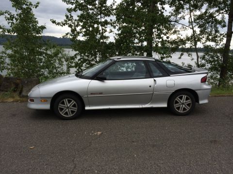 1990 Geo Storm GSi - Side view of a silver 1990 Geo Storm GSi hatchback parked outdoors near a body of water.