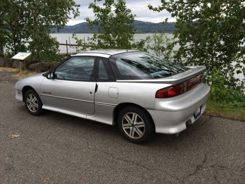 1990 Geo Storm GSi - A silver coupe is parked on a paved surface next to a body of water with trees.