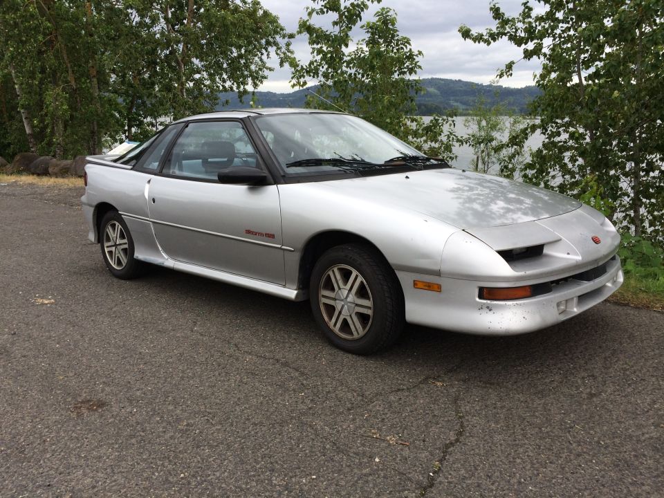 Silver 1990 Geo Storm GSi parked on asphalt near trees and water