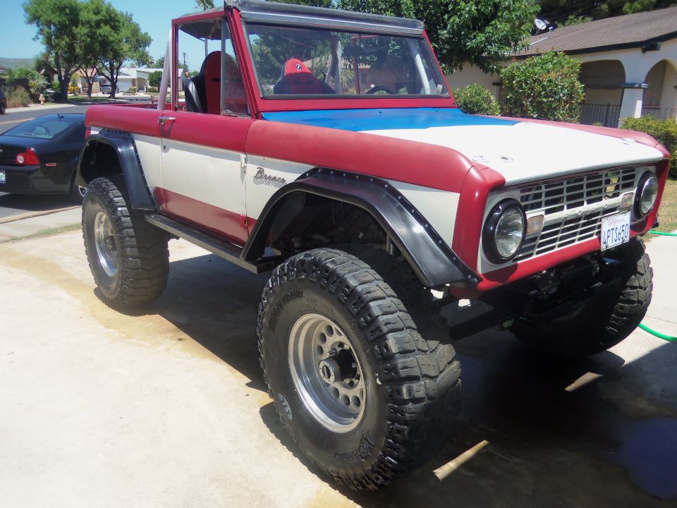 1968 Ford Bronco with red, white, and blue paint, off-road tires, and fender flares. Black car in background
