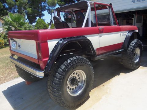 1968 Ford Bronco - A red and white 1968 Ford Bronco is shown from the rear three-quarter angle, equipped with large off-road tires.