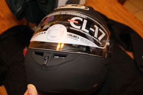 2003 Ford Crown Victoria - Rear three-quarter view of a matte black HJC CL-17 helmet showing its rear exhaust vents, with a hand at the lower edge.