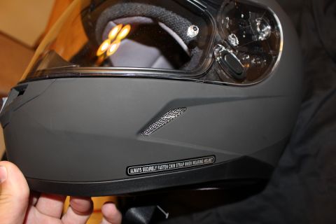 2003 Ford Crown Victoria - Detail of the lower rear of a matte black full-face helmet with a mesh vent and a sticker warning to fasten the chin strap.