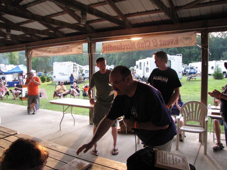 People gathered under a covered pavilion at an outdoor event with RVs and tents in the background