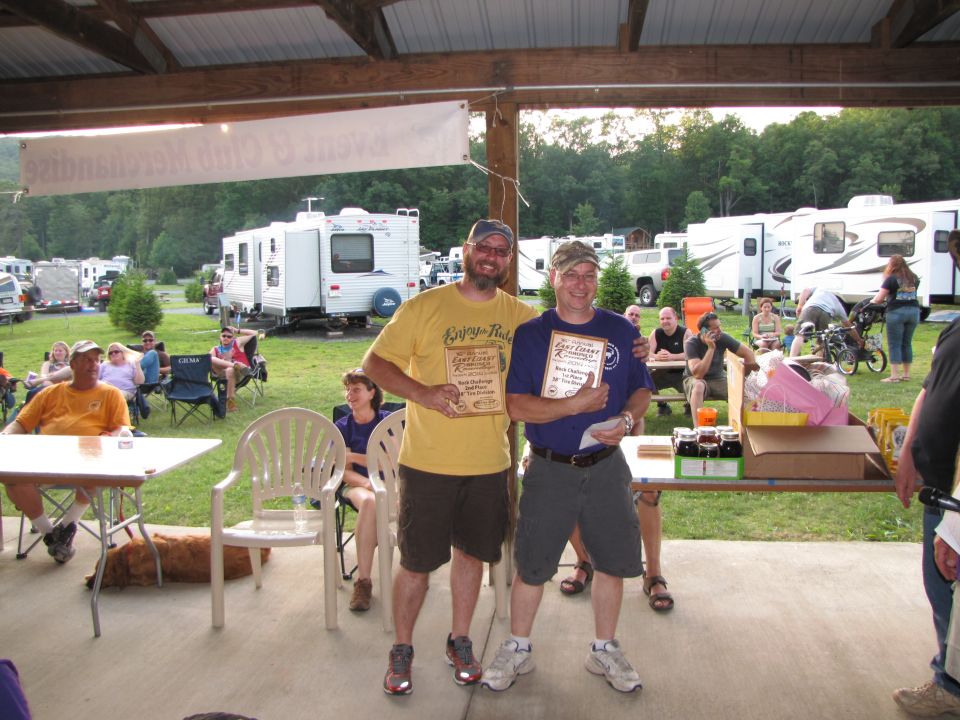 Two guys holding awards at an outdoor event with RVs in the background