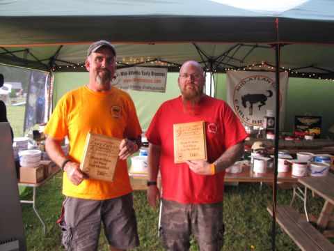 1979 Ford Bronco - Two men proudly display their awards at an event, likely a car show, with banners and tables of supplies in the background.