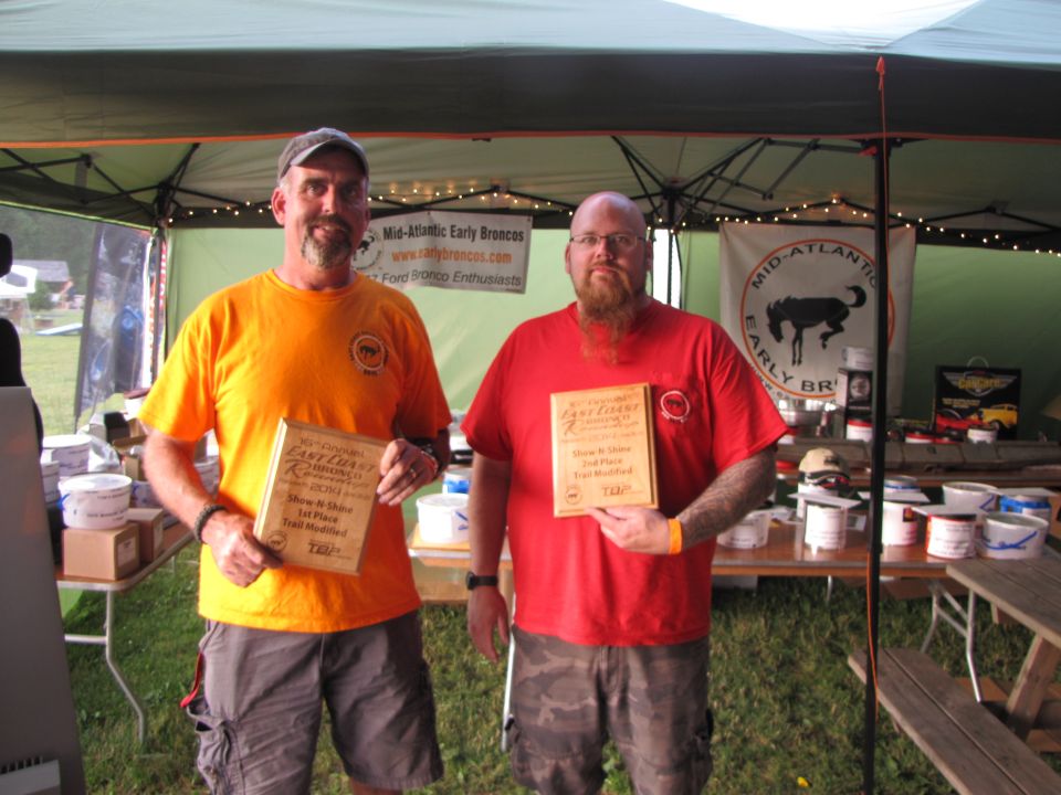 Two men stand under a tent, each holding a wooden award plaque. The man on the left wears an orange shirt and the man on the right wears a red shirt
