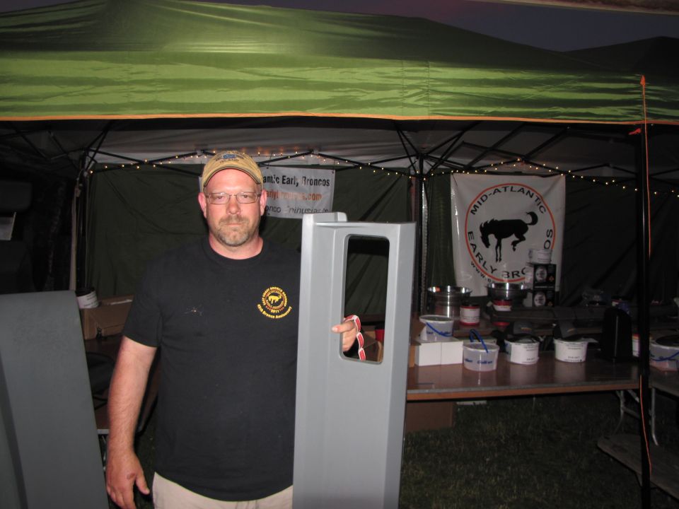 Man in cap and t-shirt holds gray car door in front of tent with banners