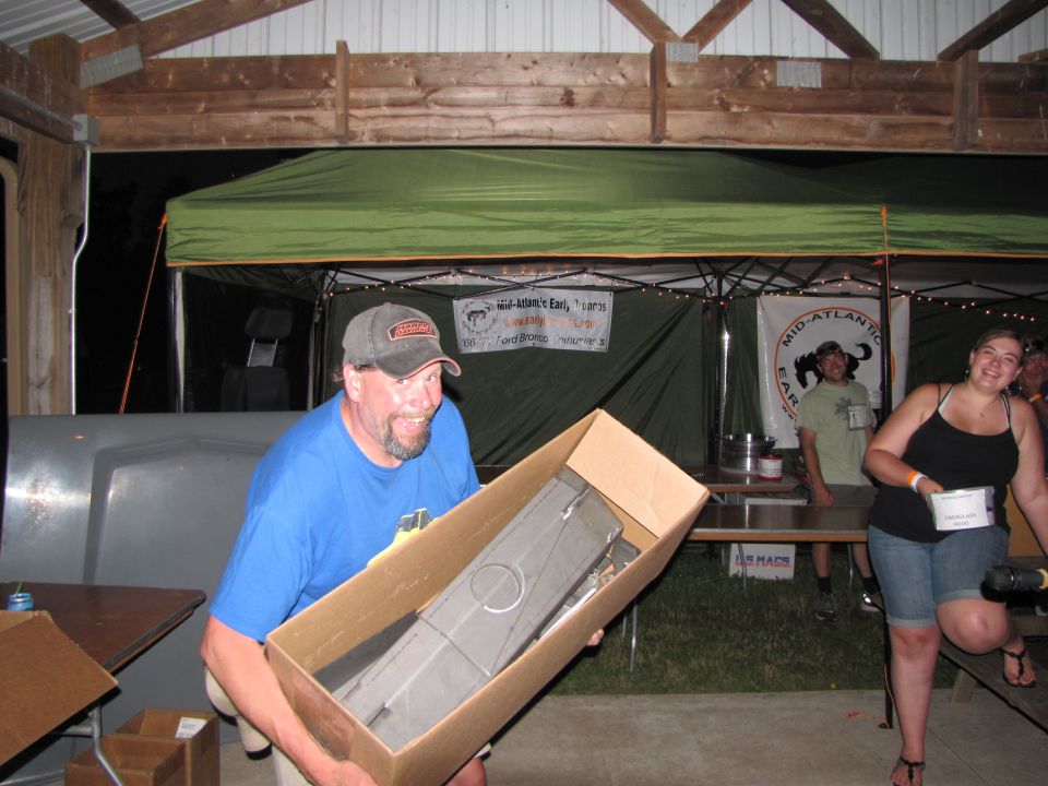 Man holds a long, gray car part in a cardboard box. Other people and tents are in the background