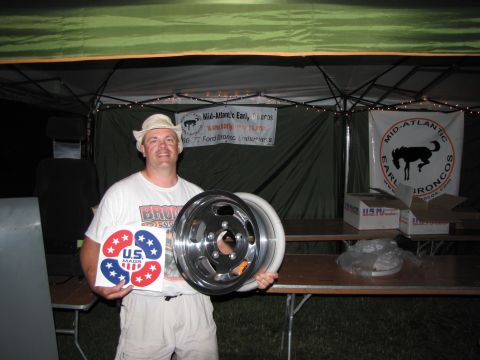 1979 Ford Bronco - A man displays a car wheel and a sticker, with event banners in the background.