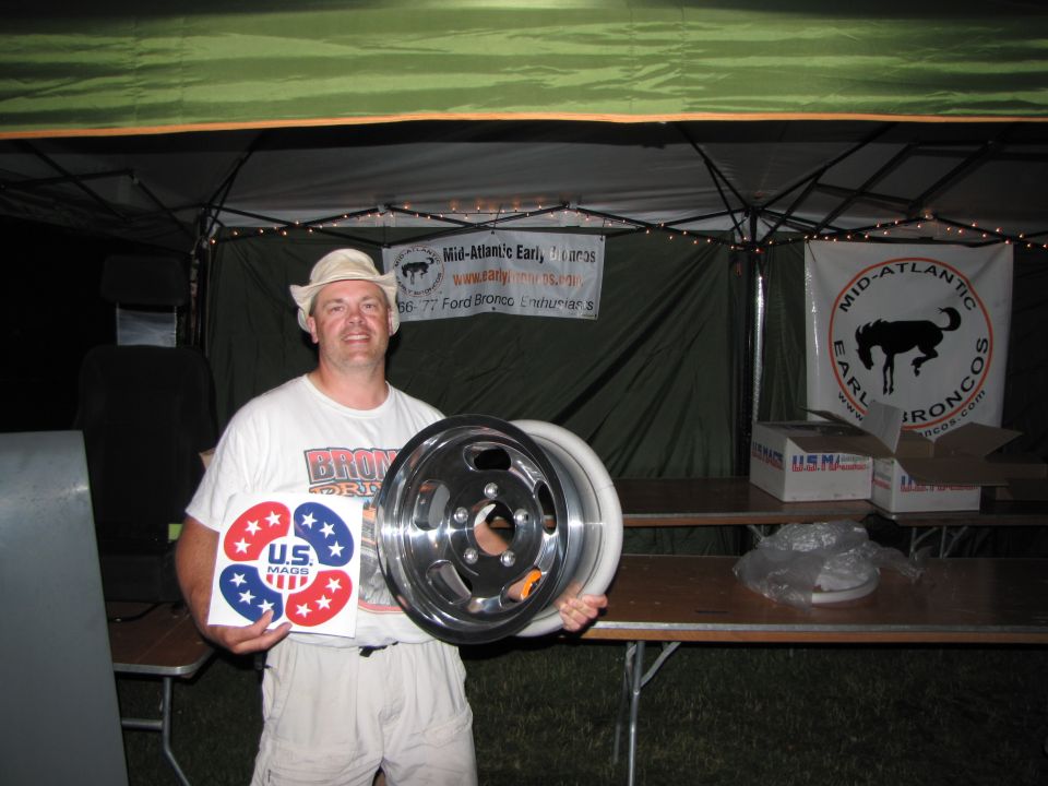 Man in hat and t-shirt holds a car wheel and a US Mags sticker. Banner behind him says Mid-Atlantic Early Broncos