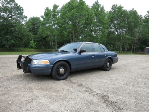2001 Ford Crown Victoria - A blue 2001 Ford Crown Victoria police car is parked on a gravel lot with trees in the background.