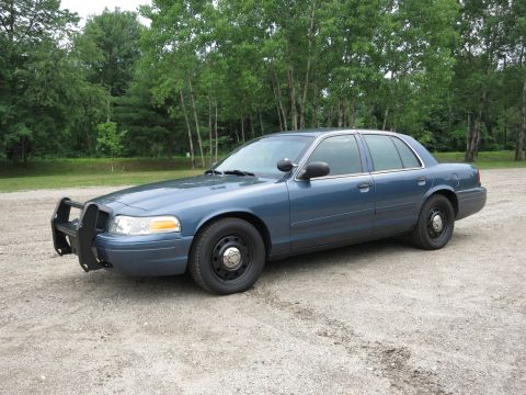 2001 Ford Crown Victoria - A blue 2001 Ford Crown Victoria police car is parked on a gravel surface with trees in the background.