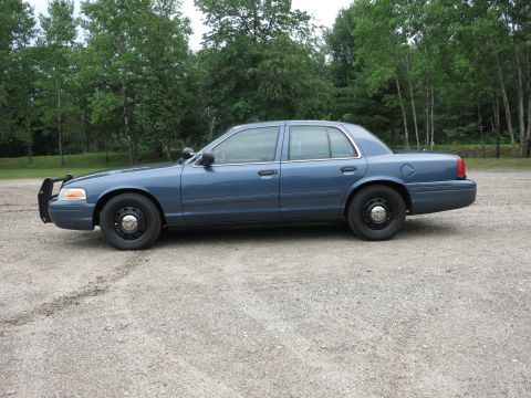 2001 Ford Crown Victoria - A blue 2001 Ford Crown Victoria sedan is parked on a gravel lot with trees in the background.
