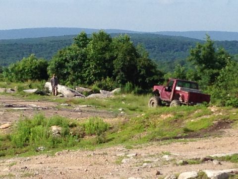 1988 Ford Bronco - A red pickup truck navigates a challenging off-road terrain with a person standing in the background.