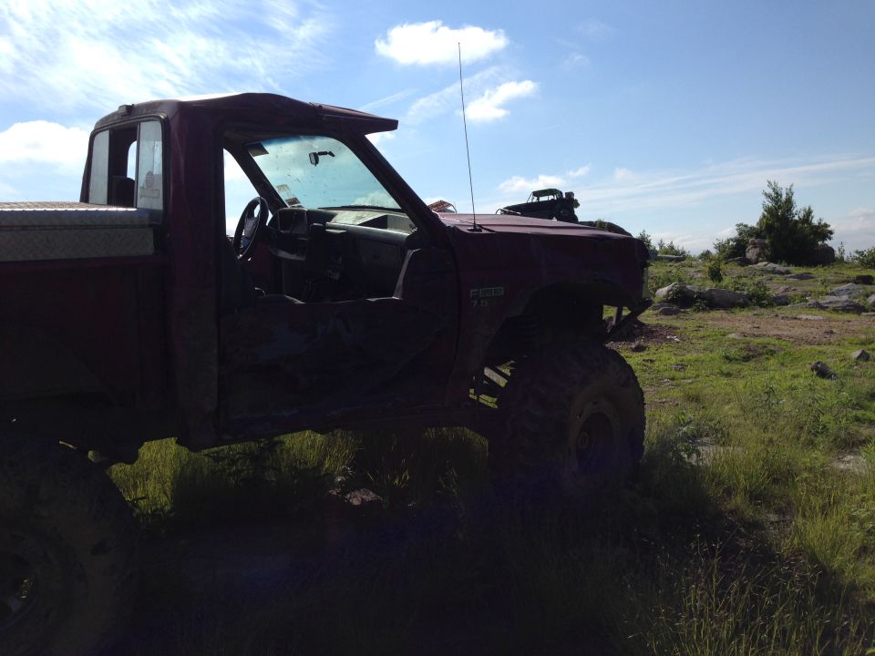 Red pickup truck with lifted suspension and off-road tires, parked on a grassy, rocky hill