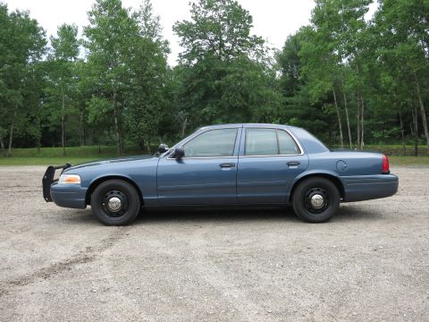 2001 Ford Crown Victoria - Side profile of a blue 2001 Ford Crown Victoria, likely a police vehicle, parked on a gravel surface with trees in the background.