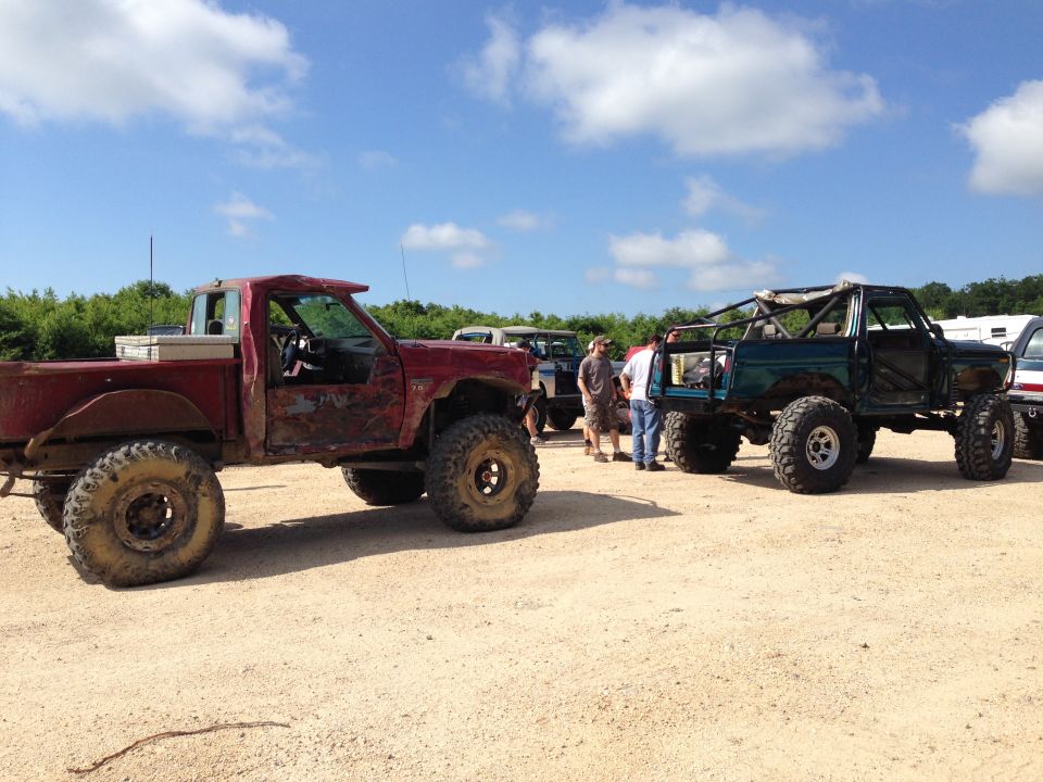 Two modified off-road trucks, one red pickup and one green SUV, parked on dirt with people around