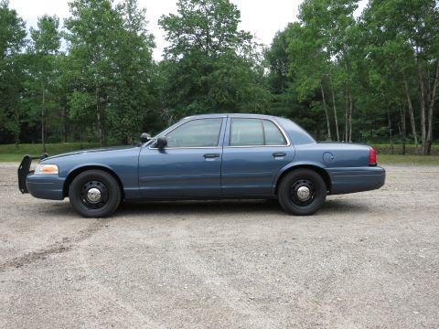 2001 Ford Crown Victoria - A blue 2001 Ford Crown Victoria sedan is parked on a gravel lot with trees in the background.