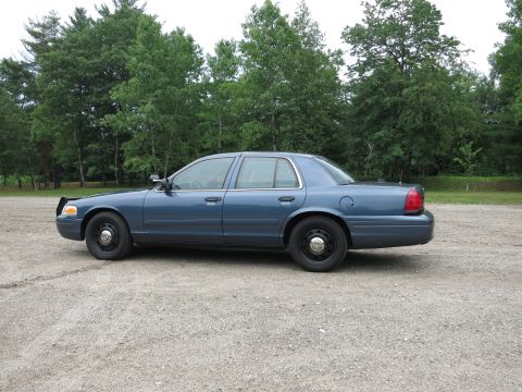2001 Ford Crown Victoria - A blue 2001 Ford Crown Victoria is shown from the side, parked on a gravel surface with a line of trees behind it.
