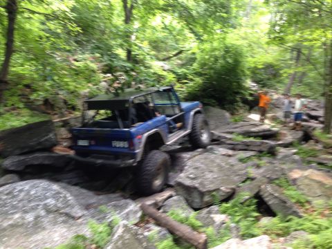 1988 Ford Bronco - A blue Ford Bronco tackles a challenging rocky terrain in a wooded area.