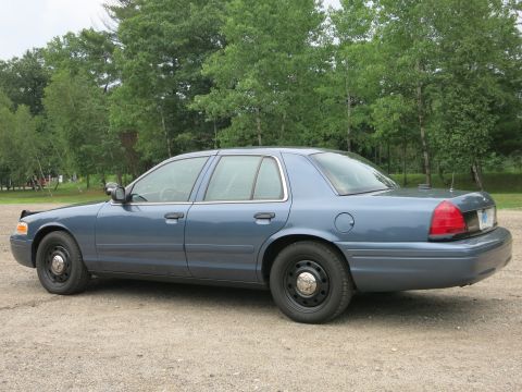 2001 Ford Crown Victoria - A blue 2001 Ford Crown Victoria is parked outdoors on a gravel surface, with a line of trees visible behind it.