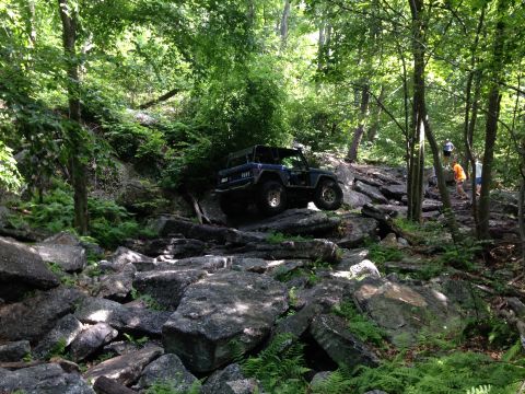 1988 Ford Bronco - A dark blue off-road vehicle tackles a challenging rocky terrain in a wooded area.